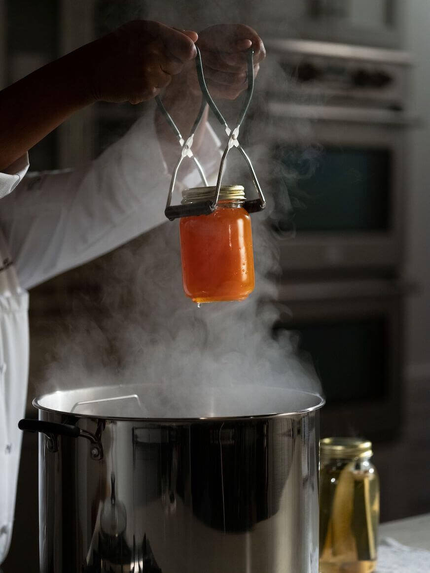 A person lifts a jar of preserves out of a steaming kettle using both hands and a pair of tongs with rubber.