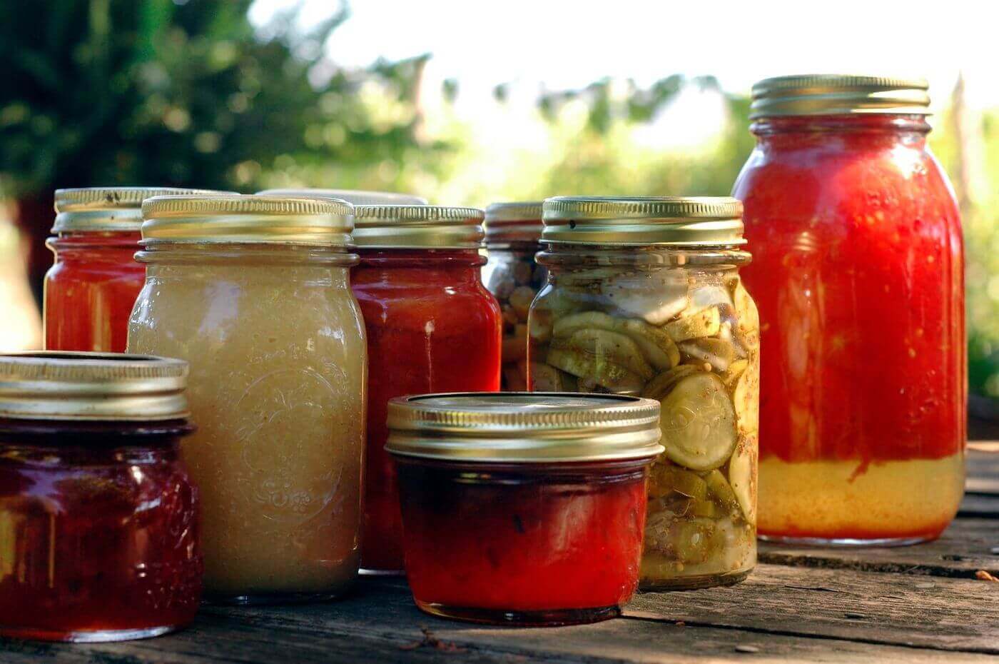 Pickles, applesauce, and preserves sit in glass jars on a rustic table outside.