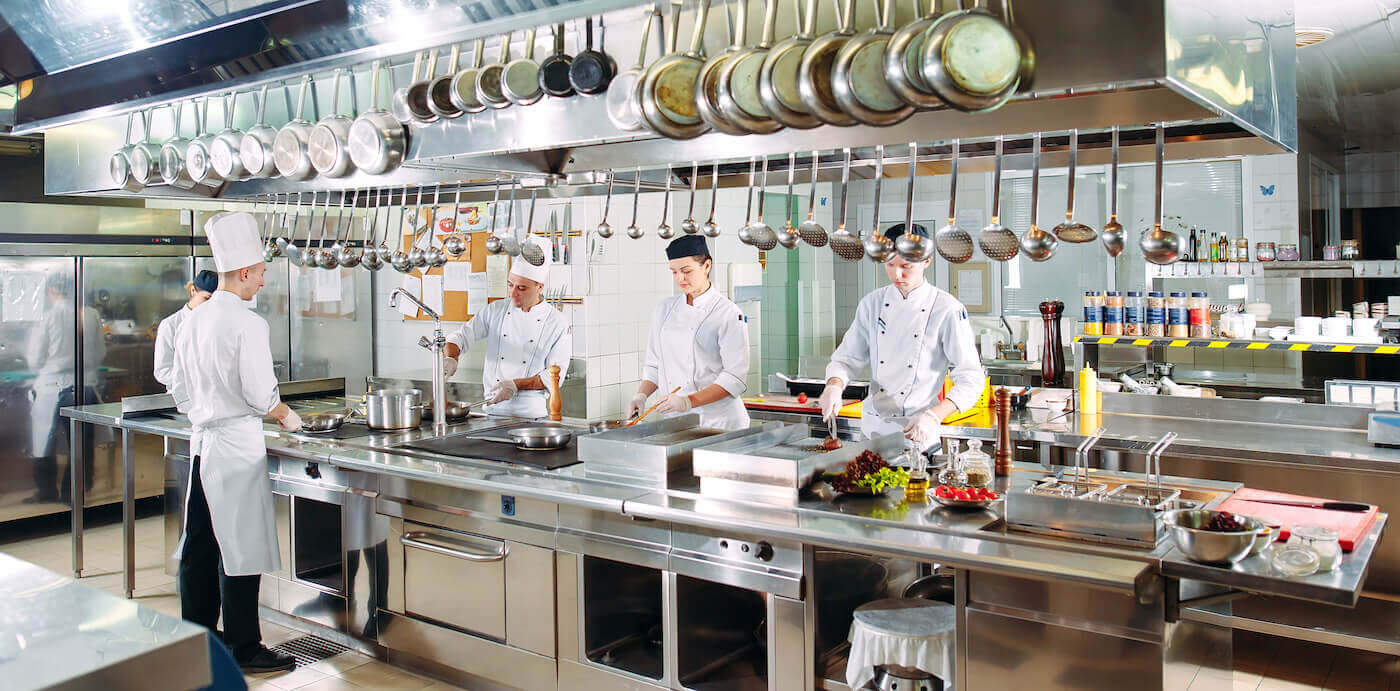 A commercial kitchen with kitchen staff at work. A number of pots and pans are hanging, and other ingredients and tools are neatly lined up.