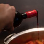 A hand pours red wine into a large kettle, which is out of focus and partially obscured.