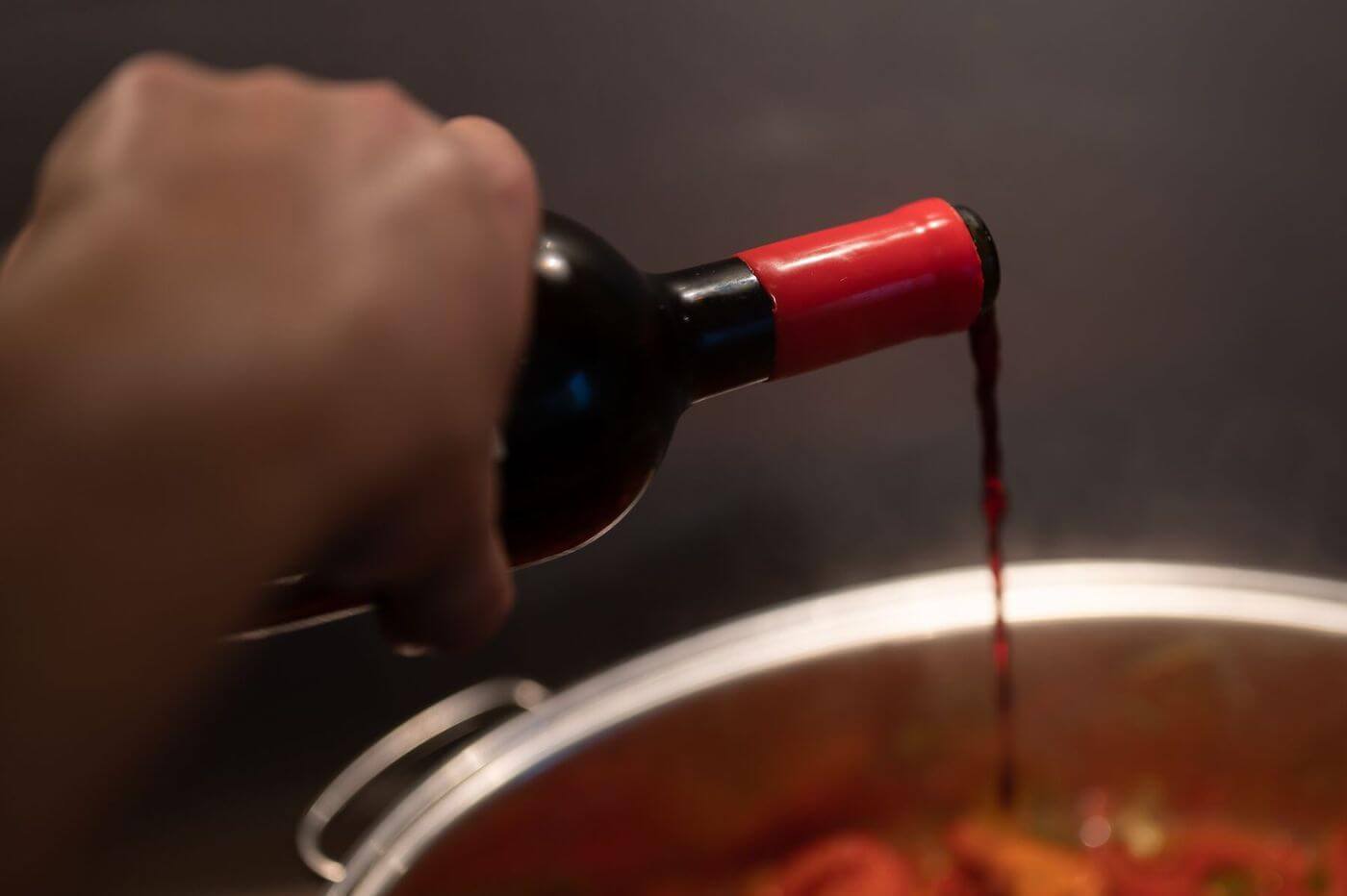 A hand pours red wine into a large kettle, which is out of focus and partially obscured.