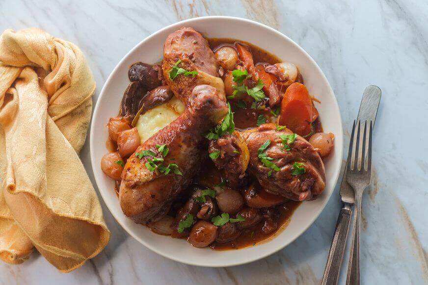 Pertunjukan coq au vin dengan jamur crimini, bawang bombay dan wortel dalam saus brendi anggur merah disajikan di atas kentang tumbuk.