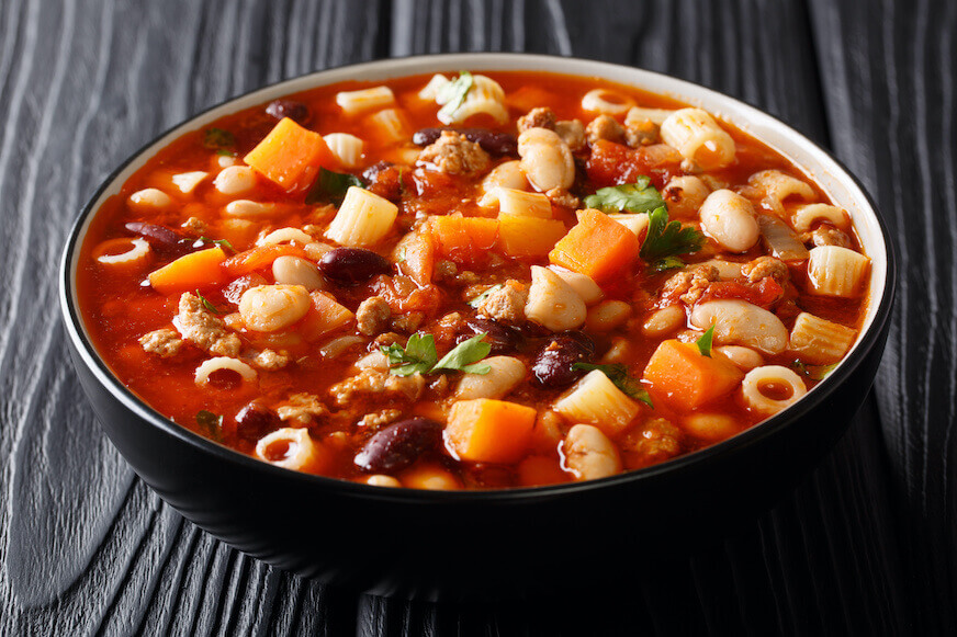 A large dish of pasta fagioli with vegetables, dinatali pasta, and ground beef, sitting on a black wooden tabletop.