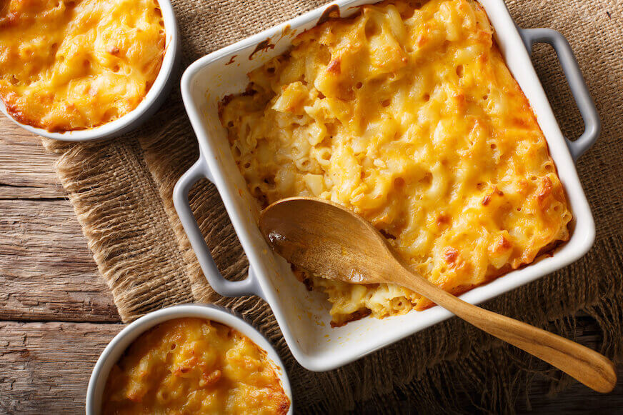 An overhead view of a square baking dish with macaroni and cheese and a wooden spoon in the dish. Two ramekins of mac and cheese sit to the side.