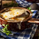 A brown crock of French onion soup with lightly browned cheese on top sits on a blue and white napkin in a rustic kitchen.