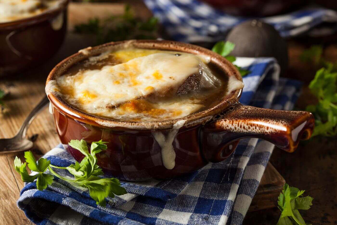 A brown crock of French onion soup with lightly browned cheese on top sits on a blue and white napkin in a rustic kitchen.