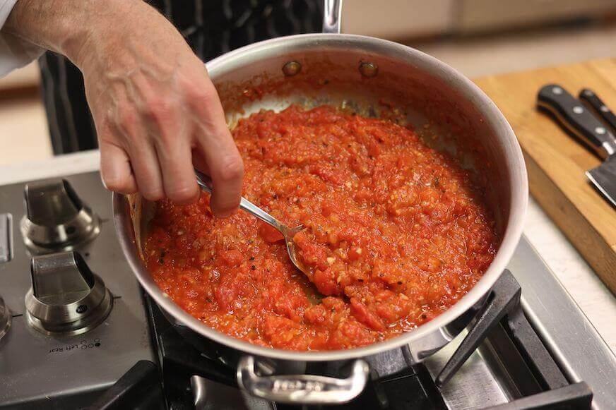 Overhead view of a stainless steel pot of tomato sauce, with a hand holding a spoon and stirring the mixture.