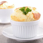 A closeup of a cheese soufflé in a white ramekin on a white saucer, with a second soufflé in the background