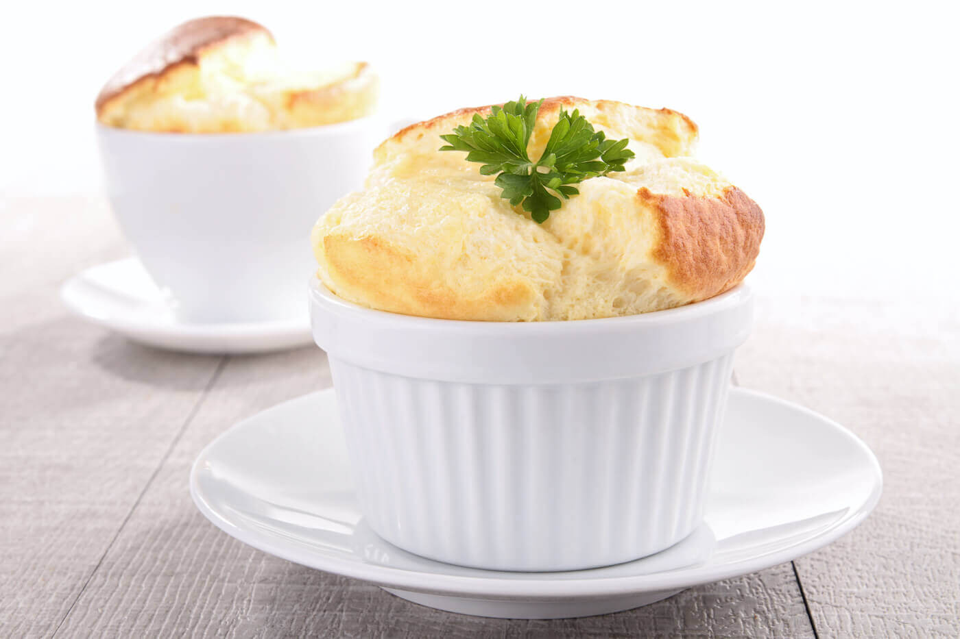 A closeup of a cheese soufflé in a white ramekin on a white saucer, with a second soufflé in the background