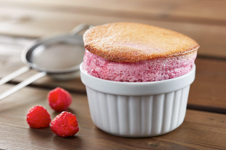 A closeup of a raspberry souffle in a white ramekin on a wooden table. A few raspberries lay nearby, and a sifter is in the background, out of focus.