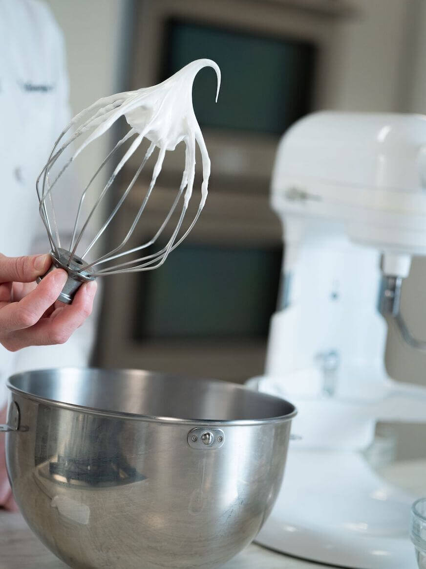 A hand holds a whisk with meringue, or whipped egg whites.