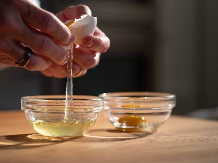 A pair of hands holds two halves of an eggshell with the yolk in one half and the whites dropping down into a clear bowl beneath them.