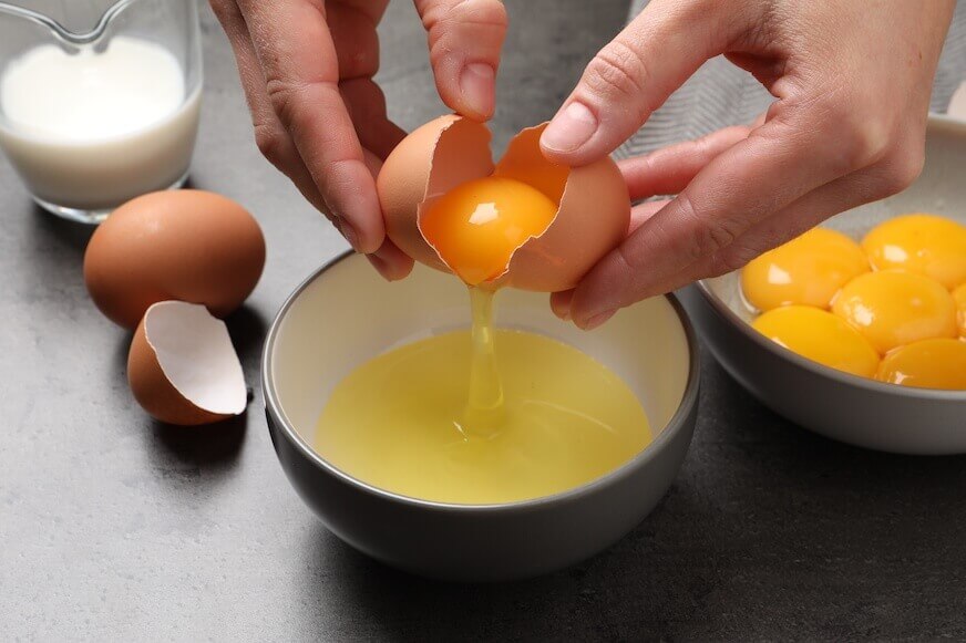 A pair of hands separating egg whites from yolks over a bowl, with a second bowl filled with egg yolks nearby.