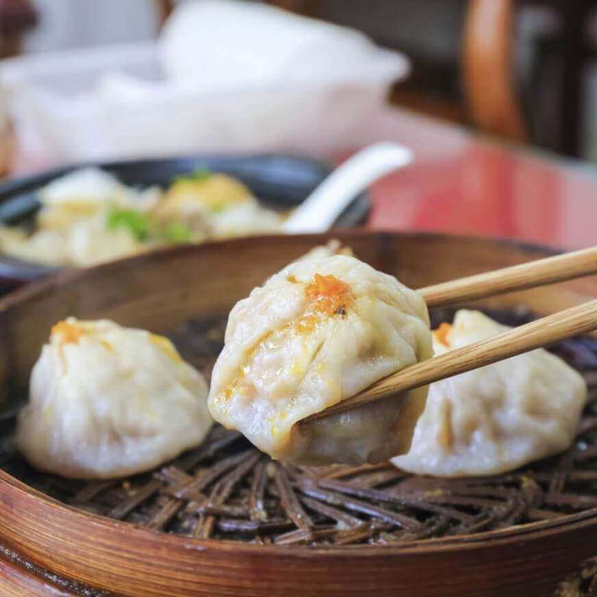 Close-up view of a dumpling being held by a pair of chopsticks. Two other dumplings sit on a bamboo basket in the background.